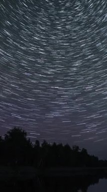 Gölün üzerindeki gece gökyüzünde yıldız izleri zaman aşımına uğradı. Sudaki yıldızların yansıması. Destansı video, dikey görüntü