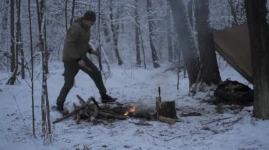 A person prepares firewood in a snowy forest, surrounded by trees and a camping tent. The cold atmosphere suggests a winter camping experience.