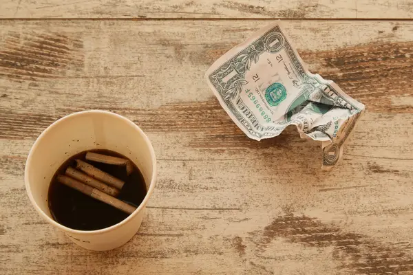 İçinde sigara izmaritleri olan bitmemiş bir kahve bardağı ve yakınlardaki masanın üzerinde buruşuk bir dolar. İş stresi