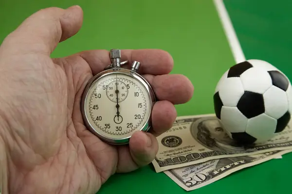 Klasik mekanik kronometre elinde ve dolar içinde yeşil arka planda futbol topu bahis oranı, bahis zamanı, spor bahisleri, bahis kazançları.  