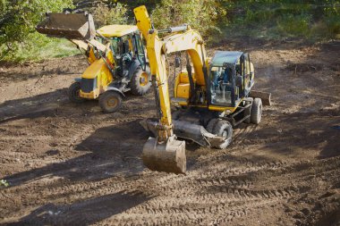 Yeşil ağaçların arasında inşaat alanında duran kazıcı ve buldozer, inşaatın ilk aşaması, dünya sahasında ağır inşaat ekipmanları