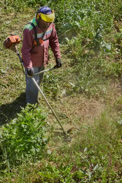 Kavurucu güneşin altında, tulumlu bir bahçıvan ve bir miğfer çim biçme makinesiyle çimleri biçiyor..