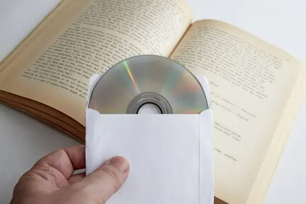 Açık bir kitabın arka planına karşılık beyaz bir zarf içinde CD tutan bir el. Metin, dijital kütüphane, eğitim kursları, kendi kendine çalışma.