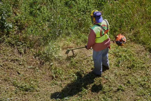 Yaz güneşinin altında, tulumlu bir bahçıvan ve miğfer, çim biçme makinesiyle uzun çimleri biçer..