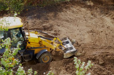 Buldozer yeşil ağaçların arasındaki inşaat sahasının zeminini düzleştiriyor, buldozer kirli bir alanda çalışıyor.