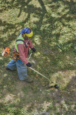 İş kıyafetleri içinde bahçıvan ve bir kask dikkatlice çim biçme makinesiyle kalın çimleri buduyor.