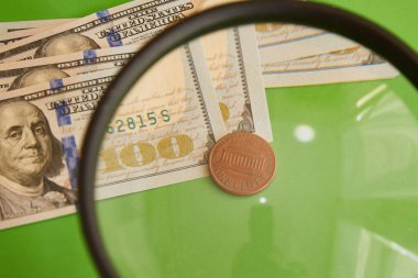 Yeşil arka plandaki büyüteçten geçen 100 dolarlık banknotların arka planına 1 sent. Dolara 1 sent. Asgari kar, asgari ücret..