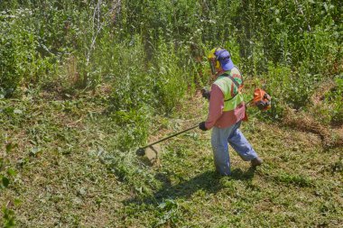 Sıcak bir yaz gününde, tulumlu bir bahçıvan ve kask manuel çim biçme makinesiyle çimlere bakar..