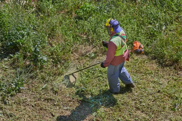 Yaz sıcağında, bir bahçıvan tulum ve kask giyer ve çim biçme makinesiyle çalışır..