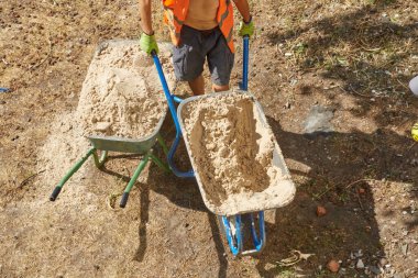 Turuncu yelek giyen bir işçi kum dolu bir el arabasını, inşaat alanında el arabası olan bir işçiyi, şantiyede ağır işçiyi itiyor..