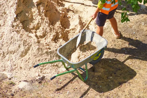 Bir işçi normal bir kürek kullanarak el arabasına kum doldurur.
