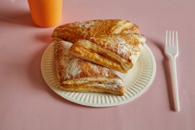 Tek kullanımlık bir tabağa şeker serpiştirilmiş üçgen puf böreği, tek kullanımlık çatal, pembe arka planda tek kullanımlık bardak, fast food, sokak yemeği..