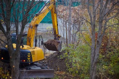 Ağaçlar, toprak fabrikaları, kazıcılar arasında inşaat alanında çalışan bir kazıcı, zor koşullarda toprağı kazarak inşaat çalışmalarına hazırlanıyor..