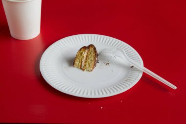  Beyaz tek kullanımlık tabakta küçük bir parça çikolatalı pasta ve kırmızı arka planda beyaz kağıt bardak, tatlı bölümü, diyet, küçük porsiyon.