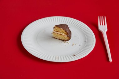 Beyaz tek kullanımlık bir tabakta küçük bir parça çikolatalı turta kırmızı arka planda beyaz bir çatal, turtadan bir pay, diyet..