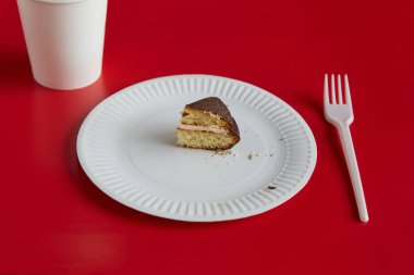 Beyaz tek kullanımlık bir tabakta küçük bir parça çikolatalı turta ve kırmızı zemin üzerinde beyaz bir kağıt bardak, bir porsiyon tatlı, diyet, küçük porsiyon.