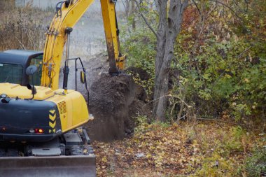 Toprakla dolu bir kazıcı kovası yere dökülüyor, sonbahar mevsiminde kazıcı işi, yakın plan, orman alanındaki bir inşaat sahasında kazı çalışması..