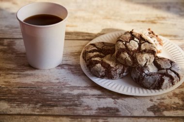 Eski bir ahşap masa üzerinde çatlaklar olan çikolatalı kurabiyelerle dolu bir kağıt tabak ve güneş ışığı demetinde bir fincan kahve, yakın çekim, kırsal kahvaltı, sade atmosfer, tatlı pişirme.