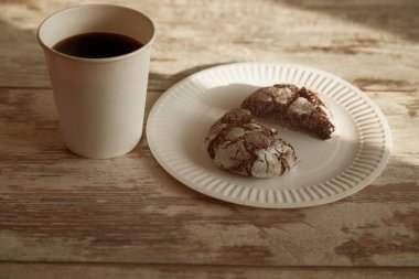 Kırık çikolatalı kurabiyenin yarısının olduğu bir tabak eski bir ahşap masa üzerinde çatlaklar ve güneş ışığı demetinde bir fincan kahve yakın çekim, kırsal kahvaltı, sade atmosfer, tatlı pişirme.