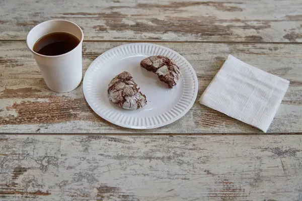 Kırık çikolatalı kurabiyenin yarısının olduğu bir kağıt tabak eski bir ahşap masa üzerinde çatlaklar ve beyaz kağıt peçetelerin yanında bir fincan kahve..