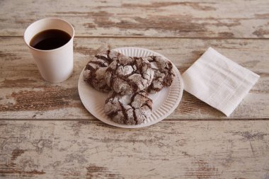 Eski bir ahşap masada çatlaklı çikolatalı kurabiyeler ve beyaz kağıt peçetelerin yanında bir fincan kahve, yakın plan, kırsal bir kafede atıştırmalık, tatlı pişirme..