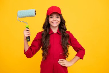 Positive and smiling builder in building uniform and cap. Painter painting with paint roller. Worker isolated on yellow background. Kids renovation concept