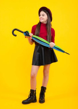 Fashion autumn kids weather, full length. Child teenager girl with umbrella in autumn skirt, sweater, boots and hat isolated on yellow studio background. Autumn trendy clothes