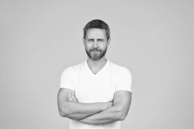 Bir erkek olarak kendinden emin olmak. Tıraşsız, kendine güvenen, gri bir geçmişi var. Beyaz adam kollarını çapraz tutuyor. Beyaz tişörtlü bir erkek. Kendine güven..