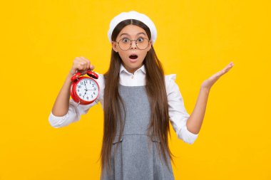 Sarı arka planda saati olan çocuk öğrenci kız. Çocuk okula geri döndü. Eğitim ve zaman kavramı. Şaşırmış yüz, şaşırmış genç kız duyguları.