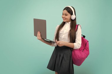 Okula dönelim. Okul üniformalı, çantalı, kulaklıklı ve dizüstü bilgisayarı olan bir kız öğrenci. İzole edilmiş mavi arka planda okul çocukları