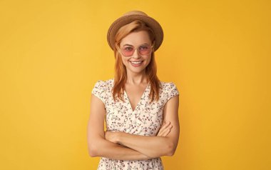 Sarı arka planda saman şapkalı ve güneş gözlüklü mutlu kadın. çapraz eller.