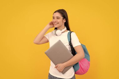 Sırt çantalı neşeli genç kız dizüstü bilgisayarı sarı arka planda kulaklıkla tutuyor, e-öğreniyor..