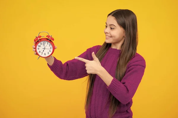 Excited face. Teenager child hold clock isolated on yellow studio background. Teenager child with alarm clock showing time. Amazed expression, cheerful and glad