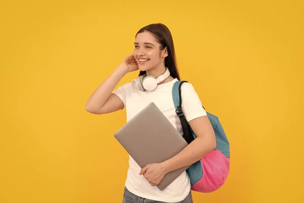 バックパックを持つ陽気な若い女の子は黄色の背景にヘッドフォンでノートパソコンを保持し ラーニング — ストック写真
