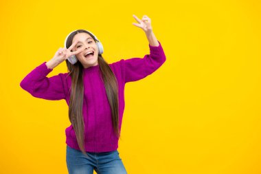 Excited face. Teenager child girl in headphones listening music, wearing stylish casual outfit isolated over yellow background. Amazed expression, cheerful and glad