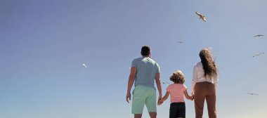 Family of mom father and child looking in future hold hands in summer, back view, summer vacation. Banner of family embracing on sea beach. Summer family vacation, horizontal banner