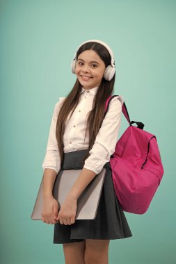 Okula dönelim. Okul üniformalı, çantalı, kulaklıklı ve dizüstü bilgisayarı olan bir kız öğrenci. İzole edilmiş mavi arka planda okul çocukları