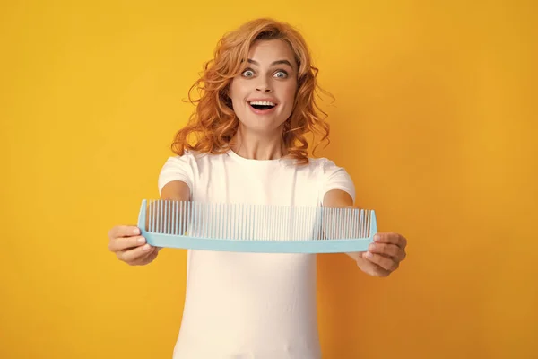 Funny excited amazed portrait of redhead woman with a comb, isolated on yellow background. Women hair care