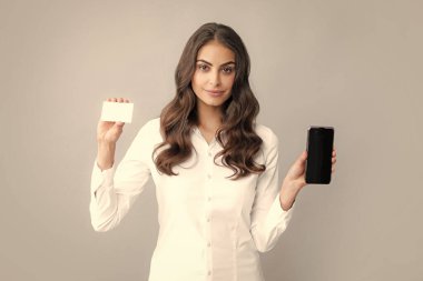Gri arka planda izole edilmiş cep telefonu kullanan kredi kartı olan mutlu genç bir kadının portresi.