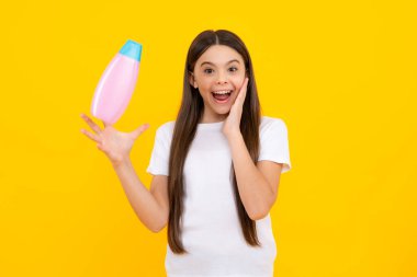 Teen child girl with shampoo bottle or shower gel isolated on yellow background. Kids hair cosmetic product. Excited teenager, glad amazed and overjoyed emotions