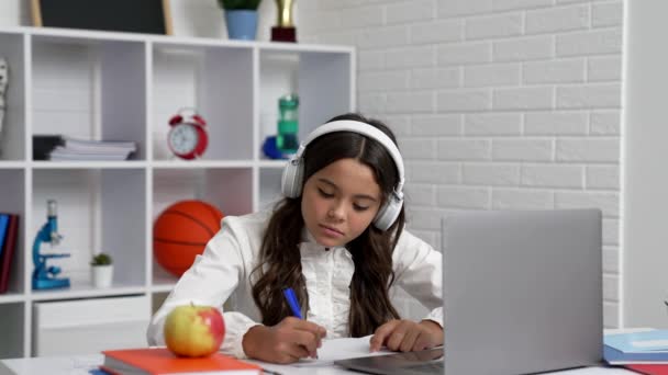 ノートパソコンで学校のオンラインレッスンで書くヘッドフォンの深刻な10代の女の子 オンライン教育 — ストック動画