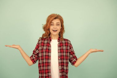 Kareli tişörtlü kızıl saçlı kadın mavi arka planda ürün sunuyor..