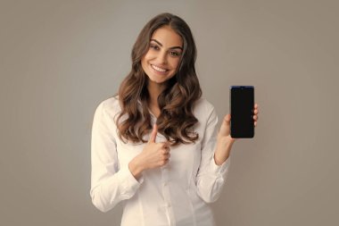 Mutlu genç kadın boş akıllı telefon ekranı gösteriyor. Tatmin olmuş, gülümseyen iş kadını cep telefonuna mesaj yazıyor, çevrimiçi iletişimden hoşlanıyor, geri bildirim türleri, gri stüdyo duvarında izole edilmiş.