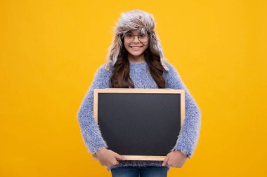 Sarı arka planda izole edilmiş, elinde karatahta tutan, sıcak bir sonbahar şapkası ve kazağı olan genç kız çocuğu. Sonbahar okulu satışı, fotokopi alanı, model