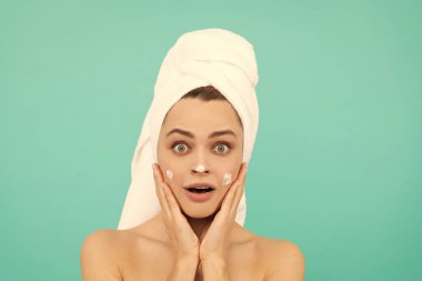Şaşırmış genç bir kadın mavi arka plana yüz kremi sürüyor..