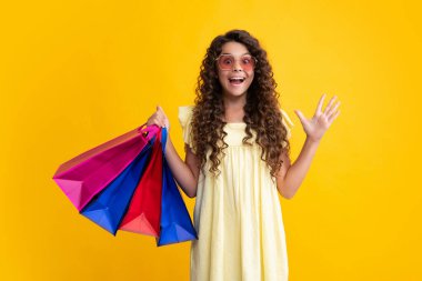 Sarı arka planda alışveriş torbaları olan güzel moda kızı. Alışveriş ve moda alışverişi. Alışveriş çantalı çocuk. Şaşırmış yüz, şaşırmış duygular.