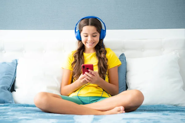 十几岁的小女孩戴着耳机 坐在房间里看智能手机上的视频 用手机笑着在网上学习的青少年学生 电子学习 — 图库照片