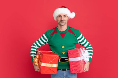 Kırmızı arka planda hediye kutusu olan xmas adamı. Mutlu yıllar. Mutlu noel hediyeleri. Elf kostümü ve Noel Baba şapkası giymiş mutlu bir adam..