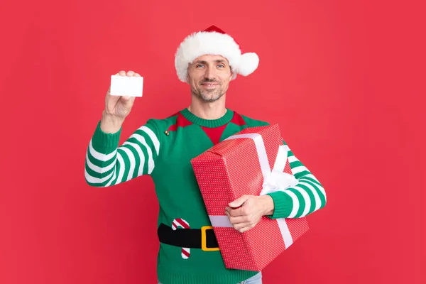 新年明けましておめでとう メリークリスマスプレゼント 買い物のためにデビットカードを持ってる男 セルフコスチュームとサンタ クラスの帽子の陽気な男 Xmas男 現在のボックスとともに赤い背景 — ストック写真