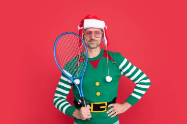 Neşeli Xmas elemanı kırmızı arka planda tenis roketi tutuyor. Mutlu yıllar. Mutlu noeller. Elf kostümlü ve Noel Baba şapkalı bir adam..